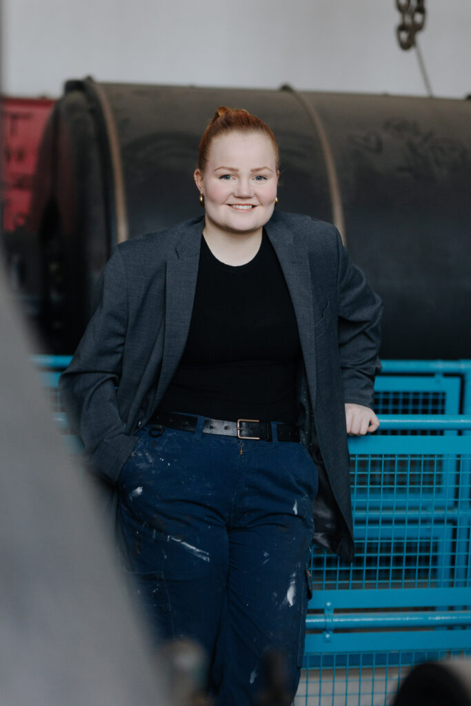 Kasketan yrittäjä katsoo suoraa kameraan, nojaten samalla vanhan puuvillatehtaan generaattorisalin kaiteeseen, päällään työhousut ja pikkutakki. 