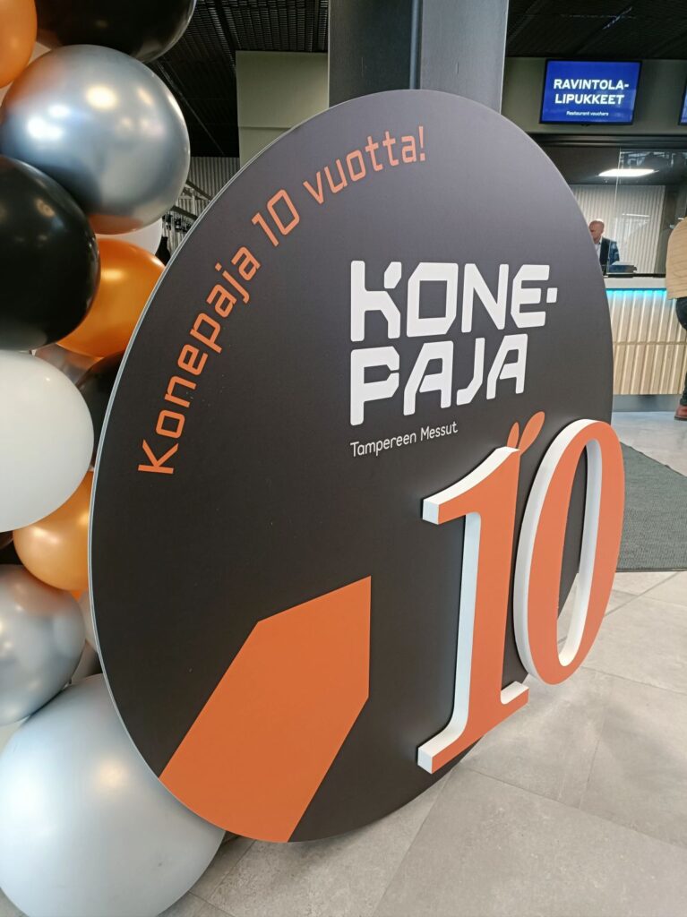 Konepaja-messut täyttivät tänä vuonna 10 vuotta, kuvassa on sen kunniaksi pystytetty pyöreä kyltti, jossa on iso numero 10 ja messujen nimi, ympärillä on kylttiin soveltuvat musta-oranssit ilmapallot.